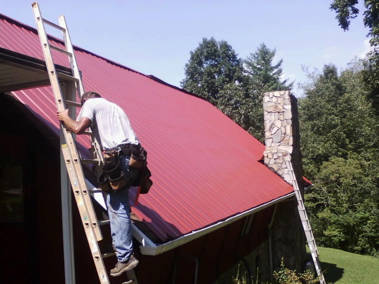 Roofer on a ladder beside a red metal roof and stone chimney — Soffit & Fascia Repair by Roofing Brothers in Fleetwood